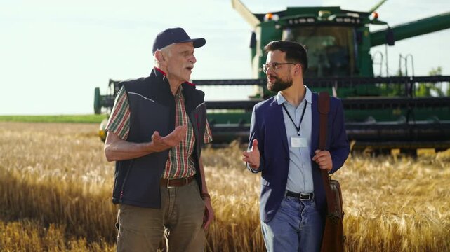 Two men are discussing farming methods in a wheat field. One man is older and shares his experience while the younger man listens and takes notes. A combine harvester is in the background.