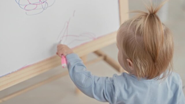 Toddler girl draws with a pink marker on a wooden standing easel in a bright home studio, hand guiding scribbles; curiosity.