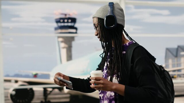 Cheerful woman walks through airport terminal with headphones on, bobbing head to music rhythm, relaxed traveler enjoying songs in audio headset.