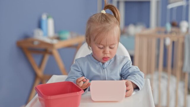 Toddler girl eating with spoon at highchair, holding spoon with hand, pink container on tray and crib in view in building; concentration curiosity.