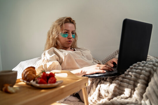 Woman with with gel eye patch working from home in bed with laptop while eating breakfast in bed 