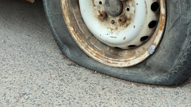 Close-up rusted rim with flat tire on asphalt road during daytime, deflated rubber bead pressed against corroded steel wheel