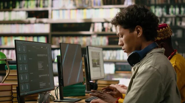 Asian student reading open book beside desktop monitor and keyboard while classmates work at nearby computers in warm library study environment, STEM education and learning, for AI learning.