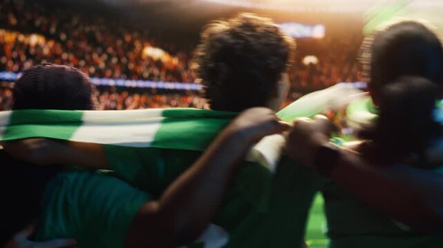 Soccer fans in stadium tribunes swaying together while watching match played on pitch, showing football supporters holding each other by shoulders during singing chant motivating players.