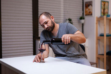 Fototapeta premium Lonely man pours wine from a bottle while drinking alone, showing depression, unhealthy alcohol use, intoxication and emotional isolation at home