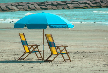 GALVESTON, TEXAS, USA: Two empty yellow canvas beach lounge chairs and a bright blue sun umbrella set up on a sandy beach on Galveston Island, looking toward the Gulf of Mexico © YuliiaK
