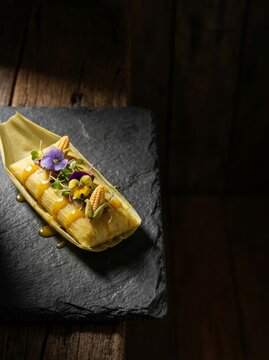 Corn tamale garnished with edible flower and sauce on slate board. Traditional mexican food dish served in husk. Gourmet cuisine presentation for restaurant menu and culinary concept.