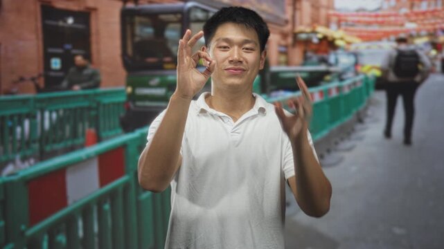 Man making ok gesture with hand on street beside green barriers and parked van, smiling in casual white polo; confidence goodwill.