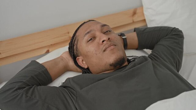 Man lying on bed with hands behind head, braids and wristwatch visible, smiling with teeth in a bedroom; contentment.