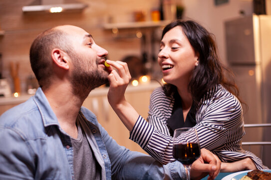 Wife feeding husband grapes during romantic dinner, wife and husband celebrating anniversary with red wine, tender moments at candle lights in kitchen.