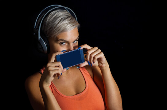 Girl whit short gray hair and headphones holding smartphone over her mouth, black background 
