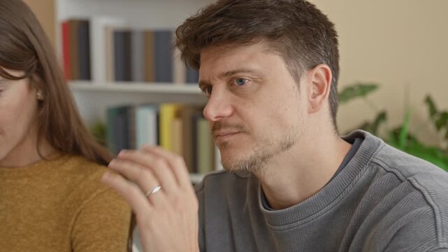 Woman in mustard sweater cups hand to mouth whispering into man's ear beside a bookshelf in a residential building; secrecy.