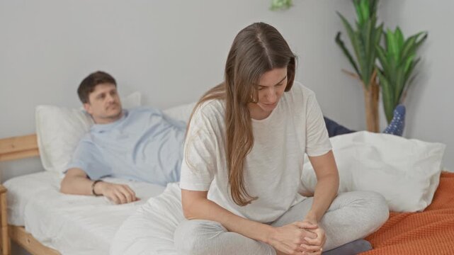 Couple on bed with woman seated crosslegged, head bowed and hands clasped while man leans in placing hand on her shoulder in a building; comfort support.