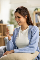 Cheerful young asian woman sitting on comfortable sofa at home with big smile holding fresh glass...