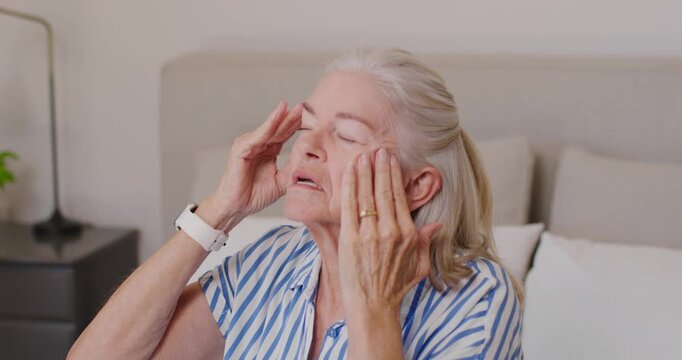 Senior woman closing eyes, pressing temples and smoothing cheeks to soothe on bed with wristwatch