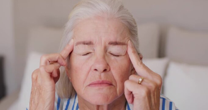 Senior female pressing temples, smoothing brow on bed after frown easing tension in shirt