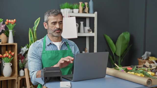 Man in green apron touches ear while studying laptop and arranging flowers in a studio; contemplation.