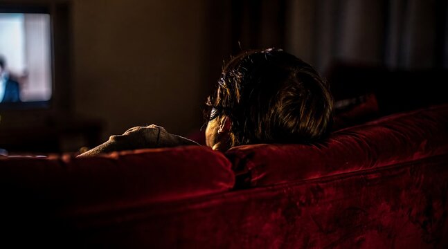 A lonely person resting their head on a red velvet couch in a dark room.