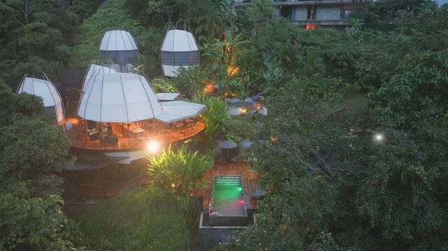 Aerial drone footage of futuristic pod-shaped eco-resort with white tent-like structures nestled in lush tropical rainforest at dusk, featuring illuminated wooden deck, glowing pool.