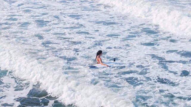 Young woman with dark ponytail wades through churning turquoise ocean waters while holding her white surfboard with mounted action camera. Bright sunlight illuminates the frothy white sea foam.