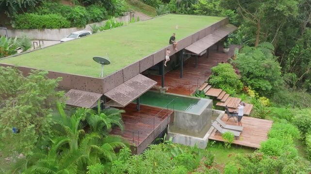 Boy jumps from roof into pool under father supervision at modern tropical villa. Aerial view captures child leaping into water while dad watches from wooden deck surrounded by lush jungle greenery.
