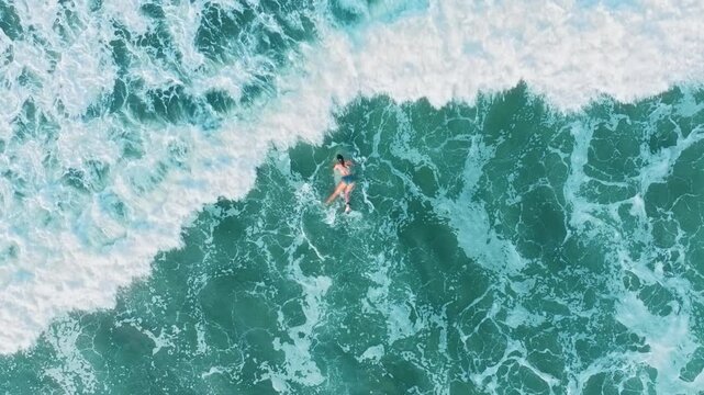 Surfing drone footage captures woman on surfboard navigating through crashing turquoise ocean waves. White foam patterns surround surfer as she paddles through vibrant blue tropical waters from above.