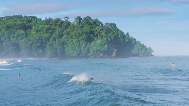 Surfing enthusiasts paddle on turquoise ocean waves near a dense tropical rainforest headland. Multiple surfers wait for swells as mist rises from the jungle-covered cliffs under a blue sky.