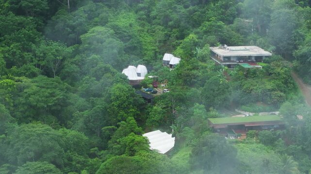 Aerial view of eco resort with geodesic dome tents and modern buildings surrounded by dense tropical rainforest. Misty fog drifts through lush green jungle canopy on hillside.