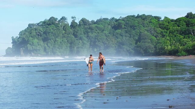 Surfing couple walks along tropical beach carrying surfboards through shallow water. Lush green rainforest covers hillside in misty morning light. Waves break on shore as surfers enjoy paradise.