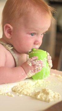 Vertical screen. Caucasian baby engages with a green sippy cup, covered in food, while seated in high chair with a messy table and playful environment