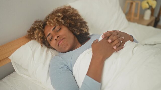 Woman lying in bed with hands clasped over chest and visible engagement ring on finger in a studio bedroom set with white bedding; quiet contemplation.