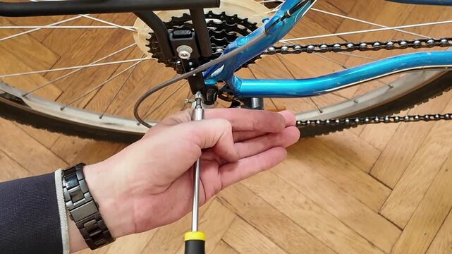 Close-up of hands adjusting bicycle derailleur with a screwdriver on a wooden floor. A person is meticulously working on a bicycle's rear derailleur system, using a screwdriver to make adjustments to 