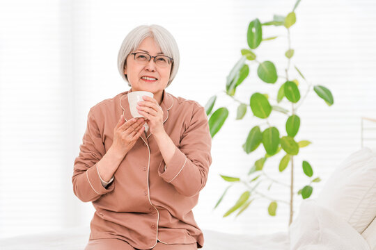 A senior woman with gray hair, wearing pajamas, rehydrates herself in her bedroom after waking up.
