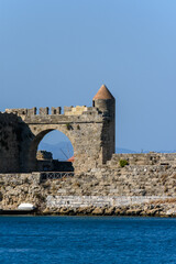 Naklejka premium Detalle de una antigua fortaleza de piedra junto al mar. Muestra un arco, una torre de vigilancia con techo cónico y aguas cristalinas en un día soleado. Ideal para turismo histórico.