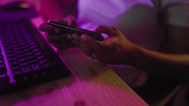 Young woman holds smartphone with both hands, thumb tapping above a computer keyboard at a studio desk lit by purple leds; focused gaming.
