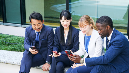 屋外ベンチでスマホを見る多国籍ビジネスパーソンのグループ © metamorworks
