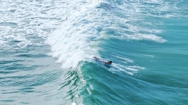 Surfing enthusiasts paddle through crystal clear turquoise ocean water as breaking waves create white foam around them. Aerial view captures two swimmers navigating powerful surf in bright daylight.