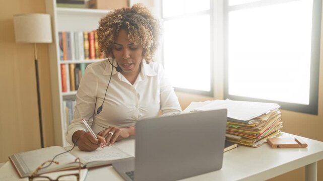 Woman writing with pen and speaking into headset at laptop near window with stacked folders in a building; concentration productivity.