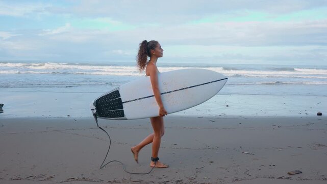 Athletic young woman with curly hair walks barefoot along wet sandy beach carrying white surfboard with ankle leash attached. Ocean waves roll in background under overcast sky.