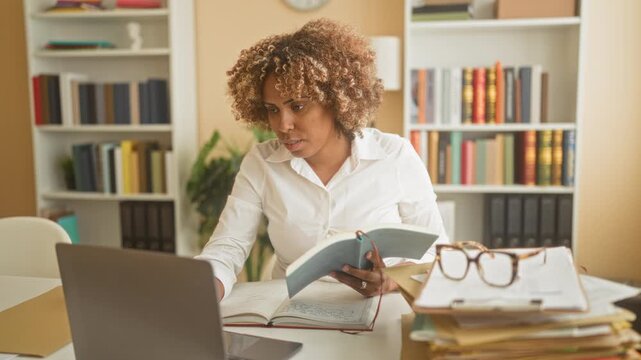 Woman holds book with left hand and uses laptop with right hand, hand on keyboard gesture in building; concentration productivity.