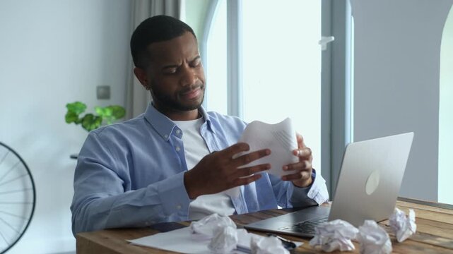 Angry African American man crumpling paper at his desk, feeling stressed and lacking ideas