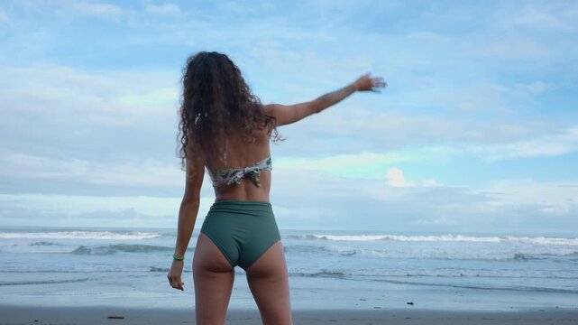 Warm up before surfing as athletic woman with curly hair performs stretching exercises on beach. She wears green bikini while practicing side bends facing ocean waves under cloudy sky.
