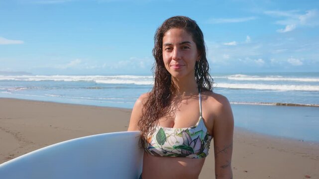 Beautiful young woman with long wet curly hair holds white surfboard on tropical sandy beach. She wears floral bikini top and smiles at camera with ocean waves and blue sky in background.