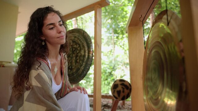 Relaxing sound meditations and sound baths session with young woman sitting peacefully near brass gong and traditional instruments in wooden outdoor pavilion surrounded by green trees and nature.