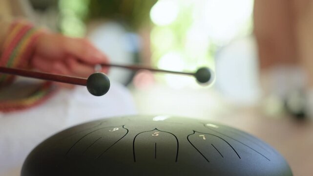 Relaxing sound meditations and sound baths therapy with green steel tongue drum. Hands holding mallets prepare to strike numbered tongues in serene natural light setting for wellness practice.