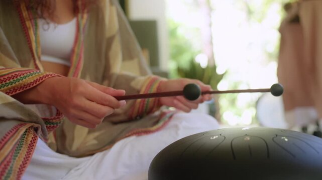 Woman wearing colorful ethnic poncho with intricate patterns holds wooden mallets over black steel tongue drum while seated in bright sunlit room with green plants and soft natural bokeh background.