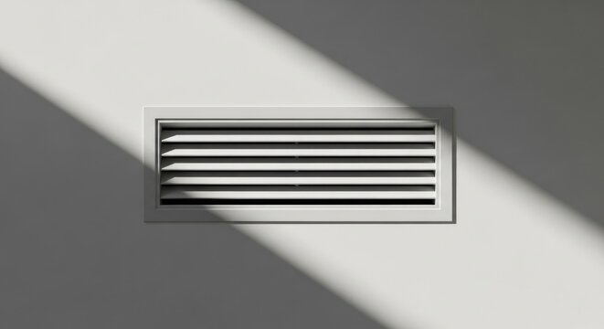 A rectangular white air ventilation vent grille mounted on a plain wall with sharp diagonal shadows cast across the surface in bright sunlight