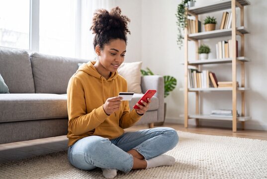 Young woman sitting on the floor at home using a smartphone and credit card for online shopping