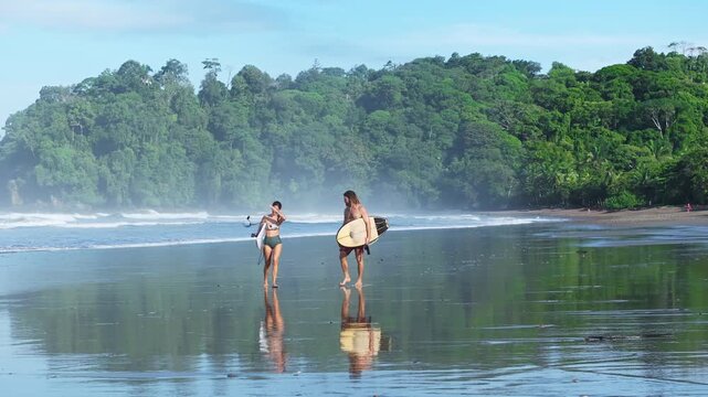 Surfing couple walks along reflective wet sand beach carrying surfboards with lush green rainforest hills in background. Misty ocean waves and tropical paradise setting create serene coastal scene.