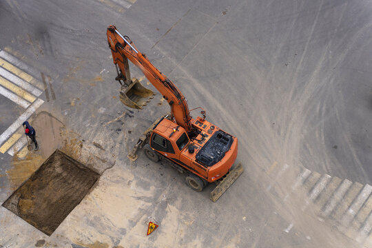 Top view of an orange wheeled excavator with raised bucket standing next to a square trench pit with a manual worker during road repair.
